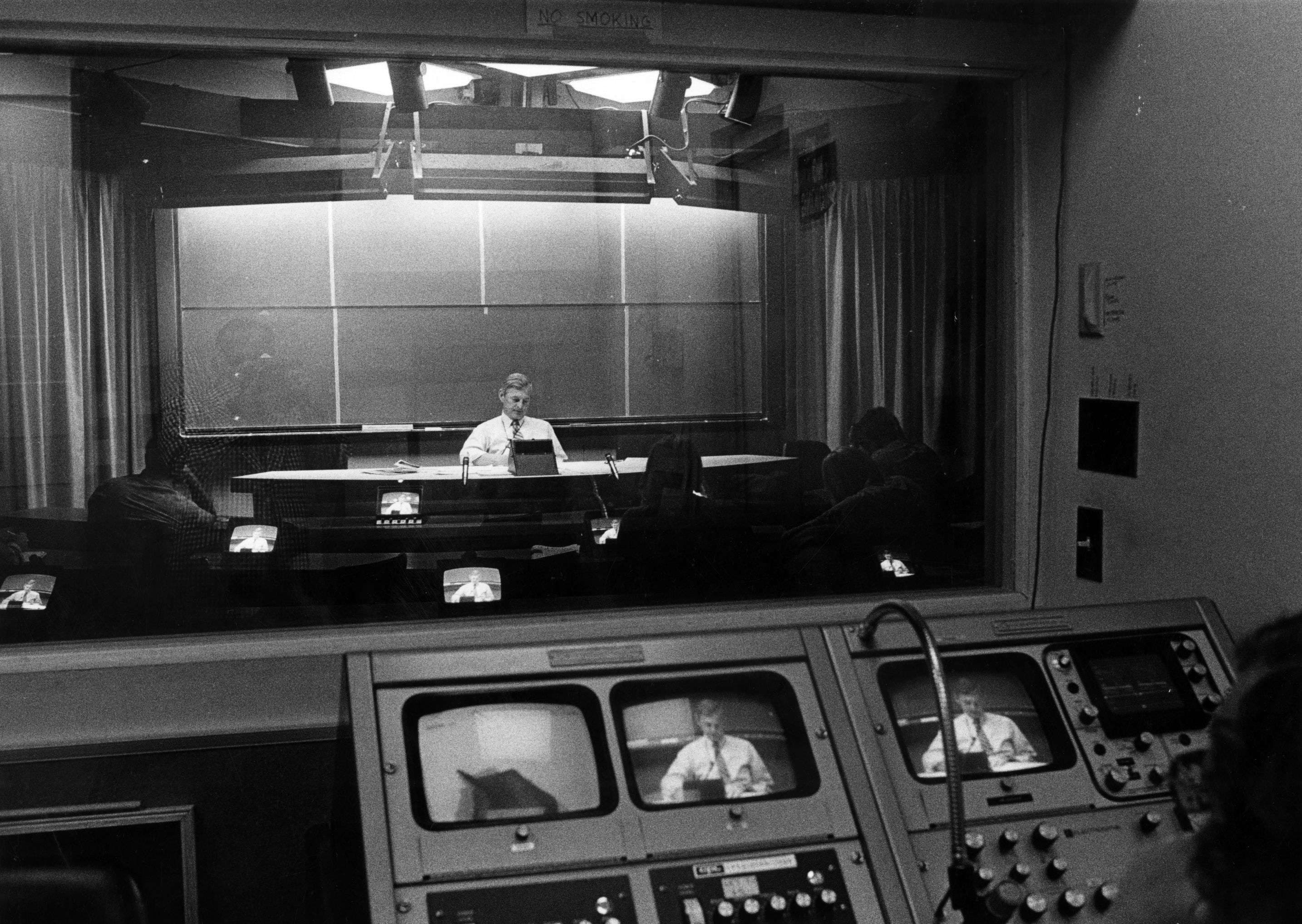A black and white image of a man surrounded by multiple monitors in a dimly lit room with onlookers in front of him.
