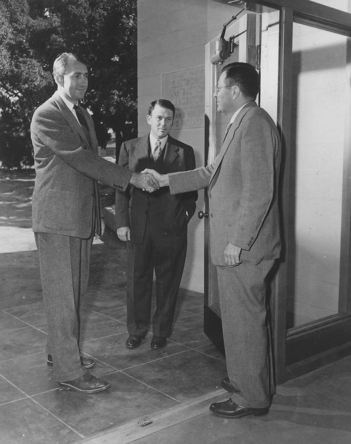 David Packard and Fred Terman shake hands at the entrance of a building while a third unnamed man stands in the background.