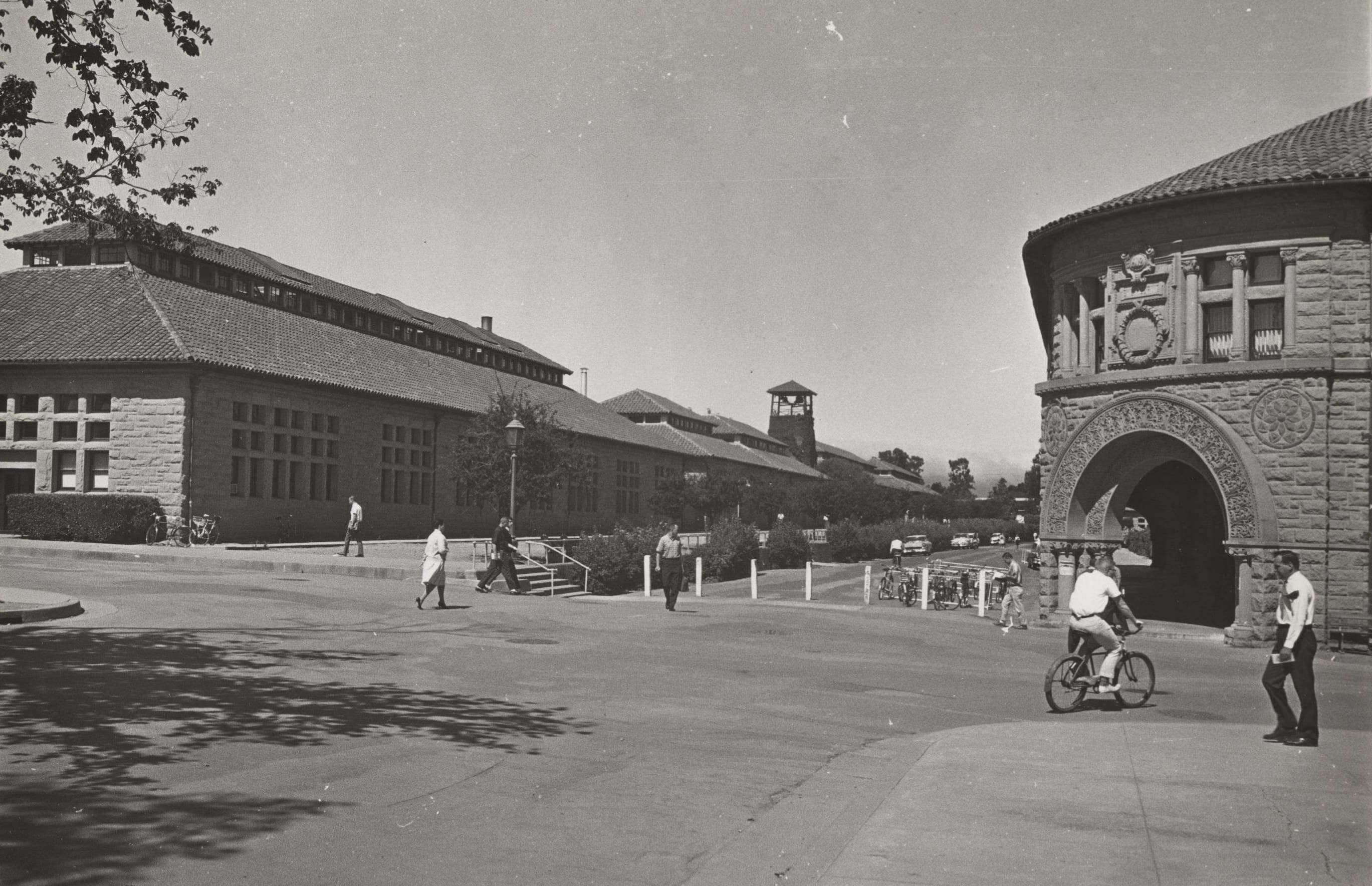Black and white photograph of individuals on bicycles in front of a building with a clock tower.
