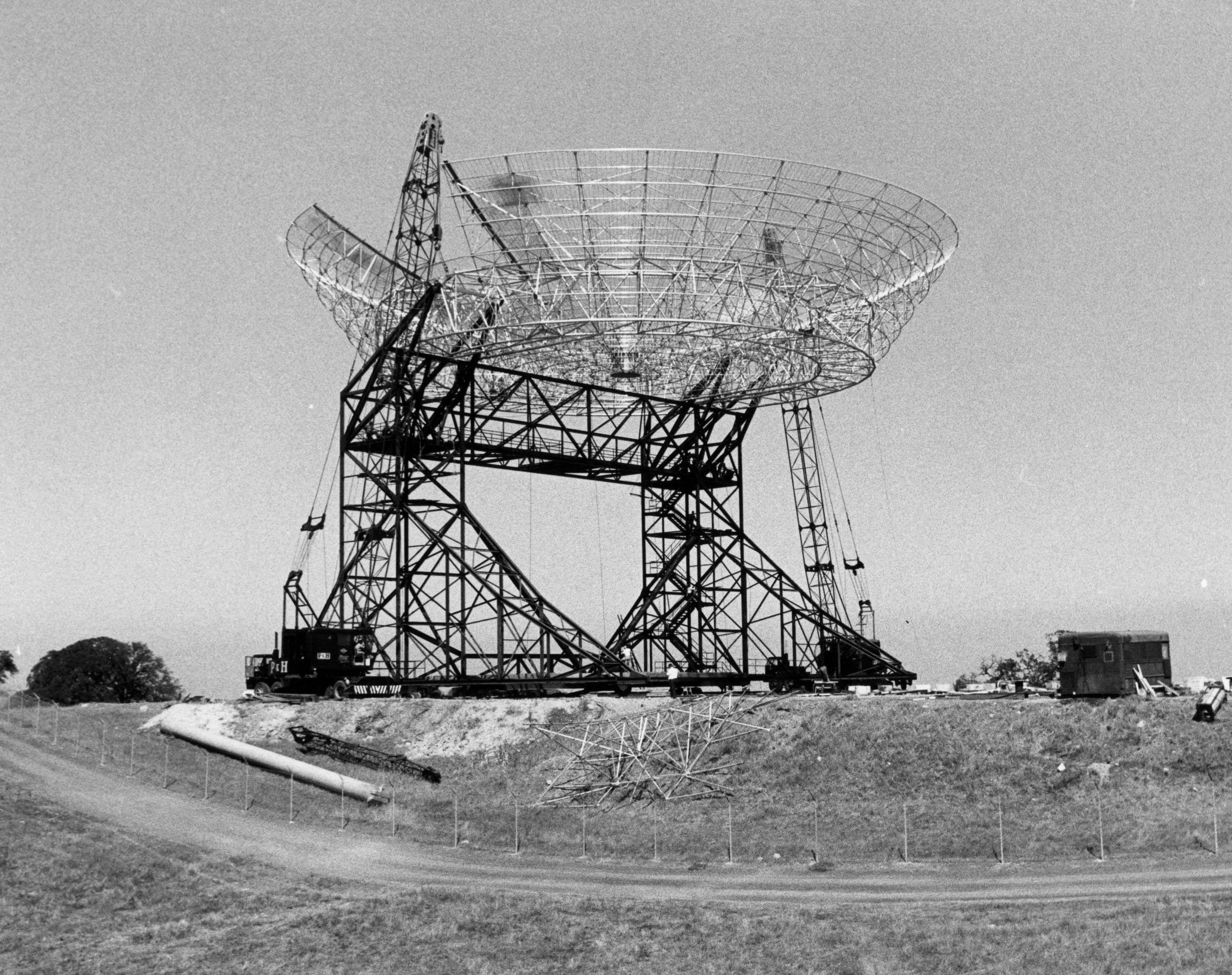 Large radio telescope dish under construction on a grassy hill, surrounded by scaffolding and construction equipment, with a clear sky in the background.