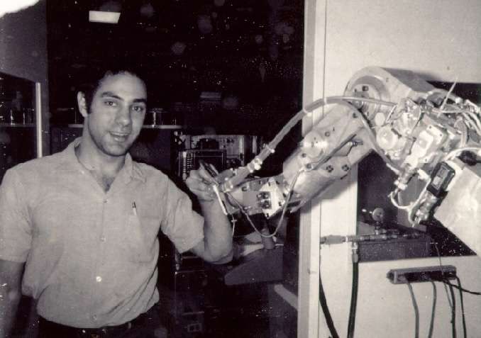 A man stands in front of a computer, holding a robotic arm.
