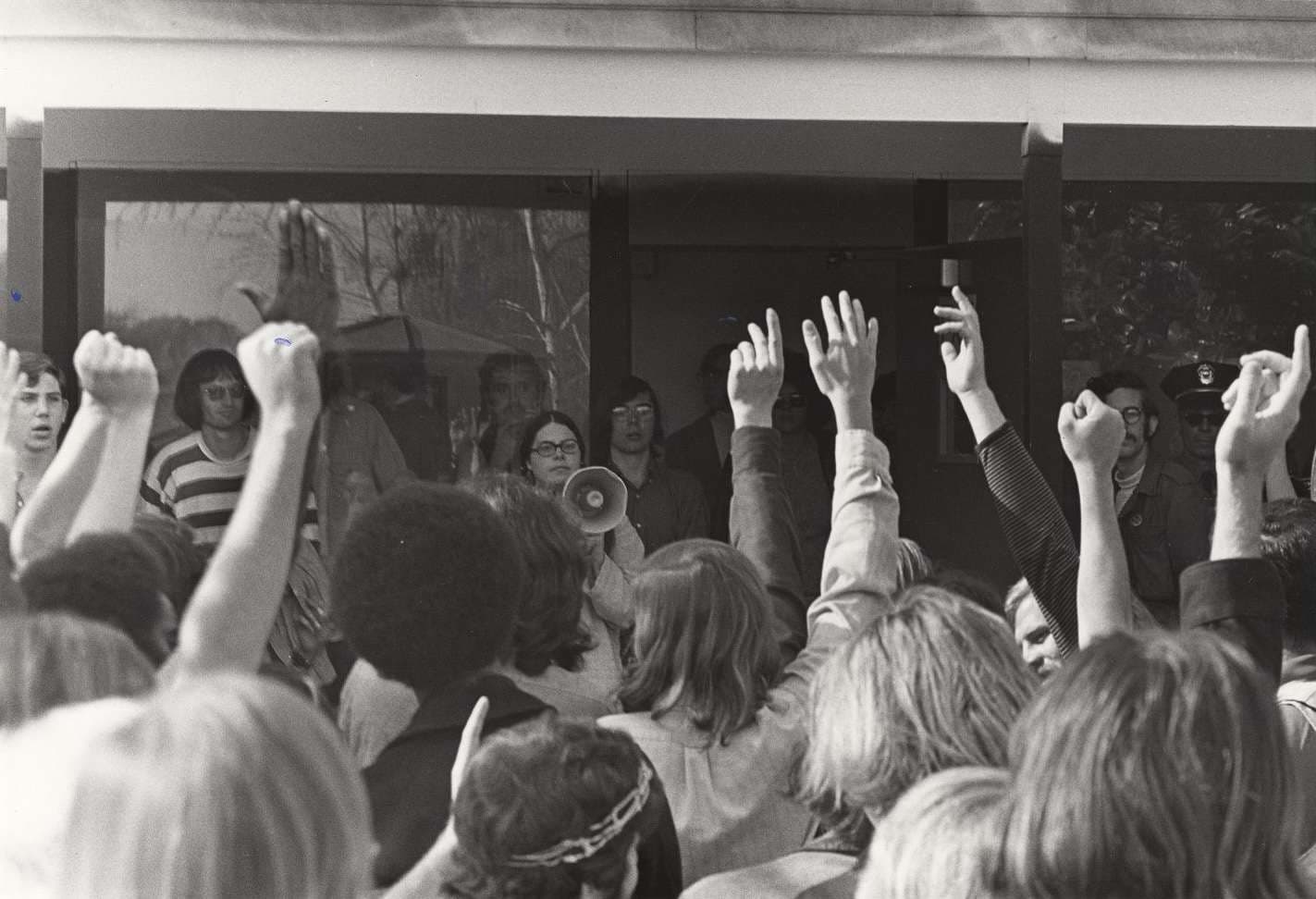 A diverse crowd gathered in front of a building, with their hands raised, indicating support in protest.