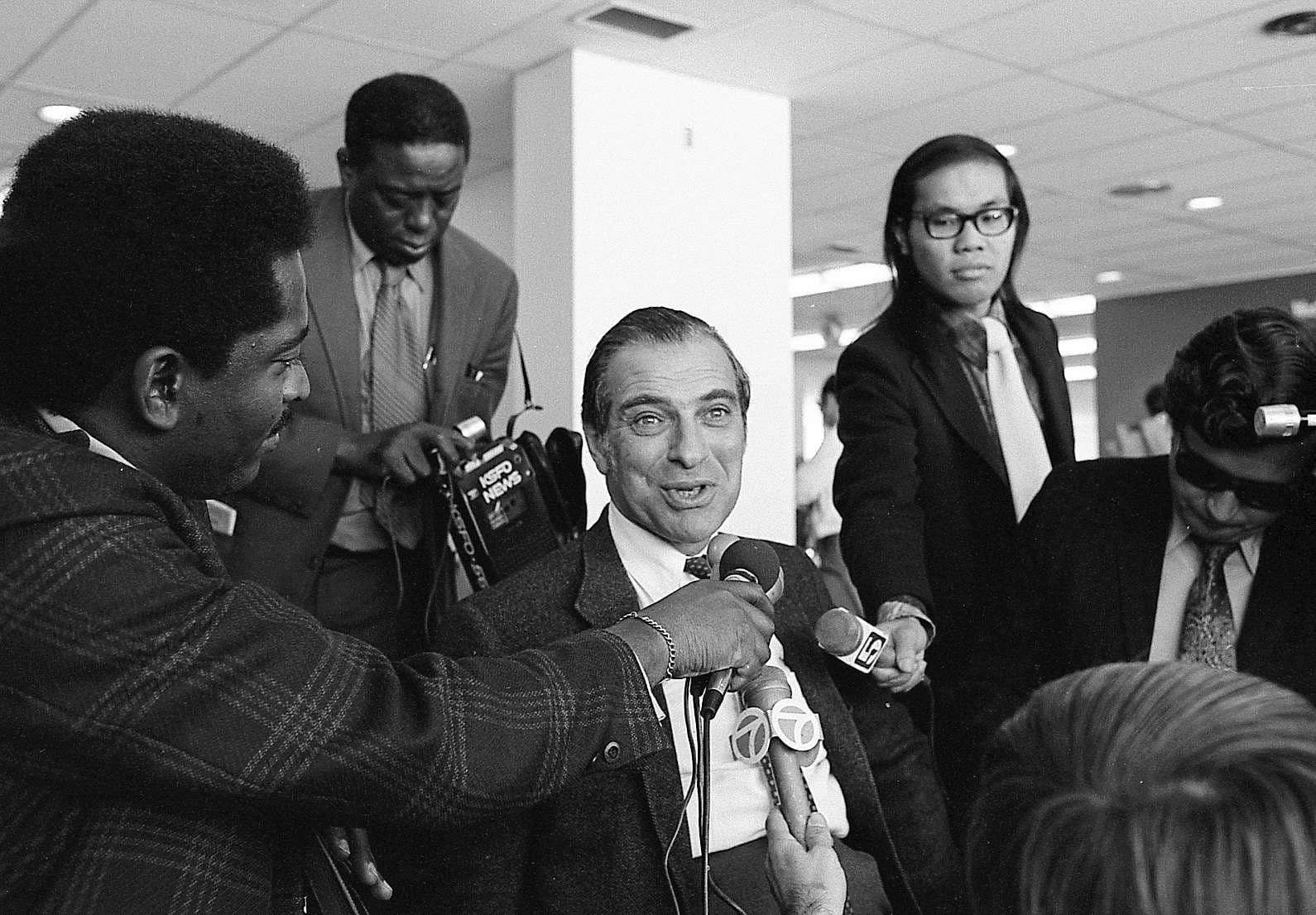 A man in a suit and tie stands confidently as reporters gather around him, capturing the moment with their cameras.