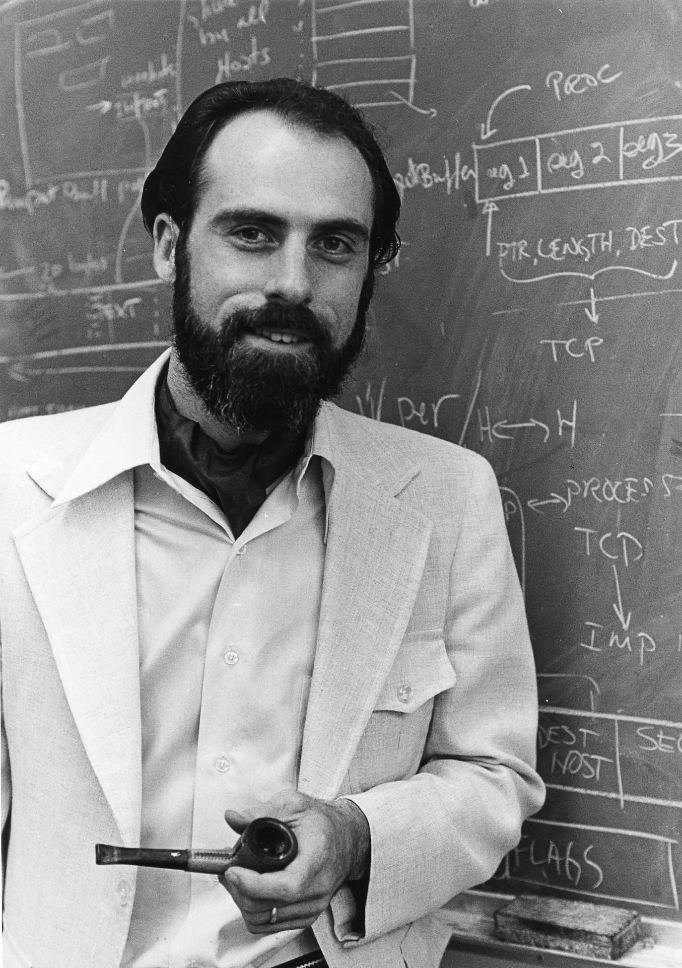 A bearded man stands confidently in front of a chalkboard holding a pipe.