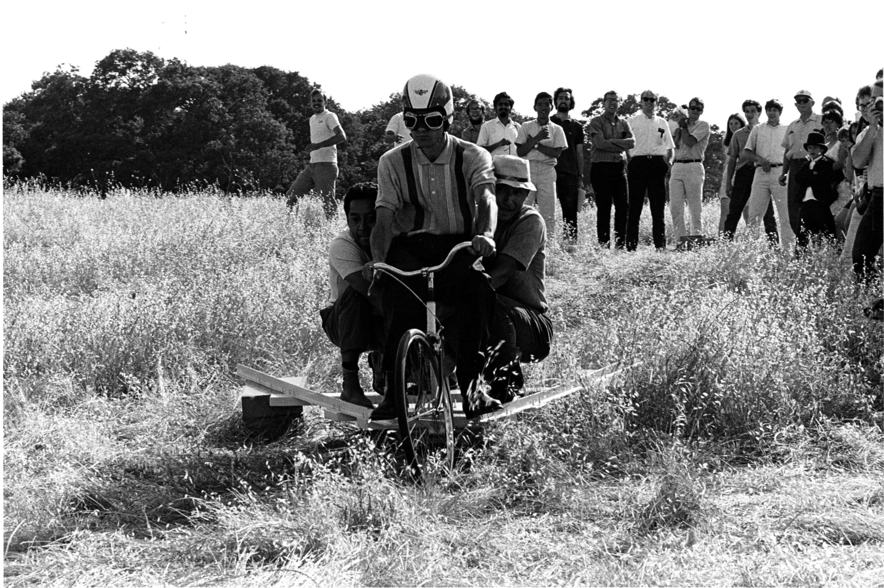 Four men ride a homemade cart downhill through a grassy field, wearing helmets, smiling, and exuding excitement. A crowd watches in the background.