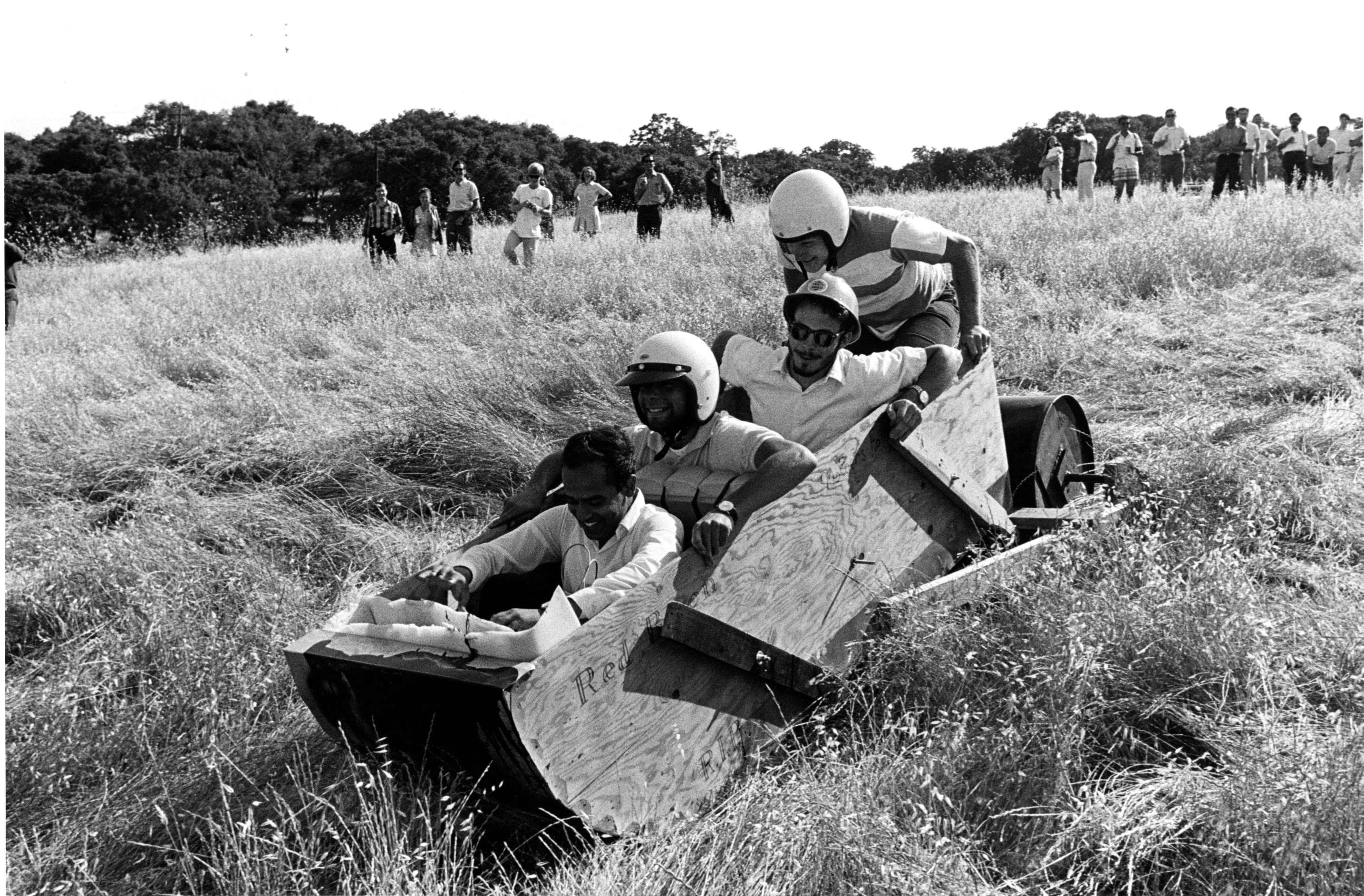 Black-and-white image of a man wearing a helmet and goggles riding a bike over a makeshift ramp. A crowd watches in a grassy field, creating a lively atmosphere.