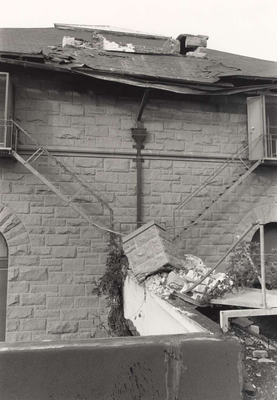 Black and white image of a building featuring a broken roof, where fragments have fallen onto a different part of the structure.