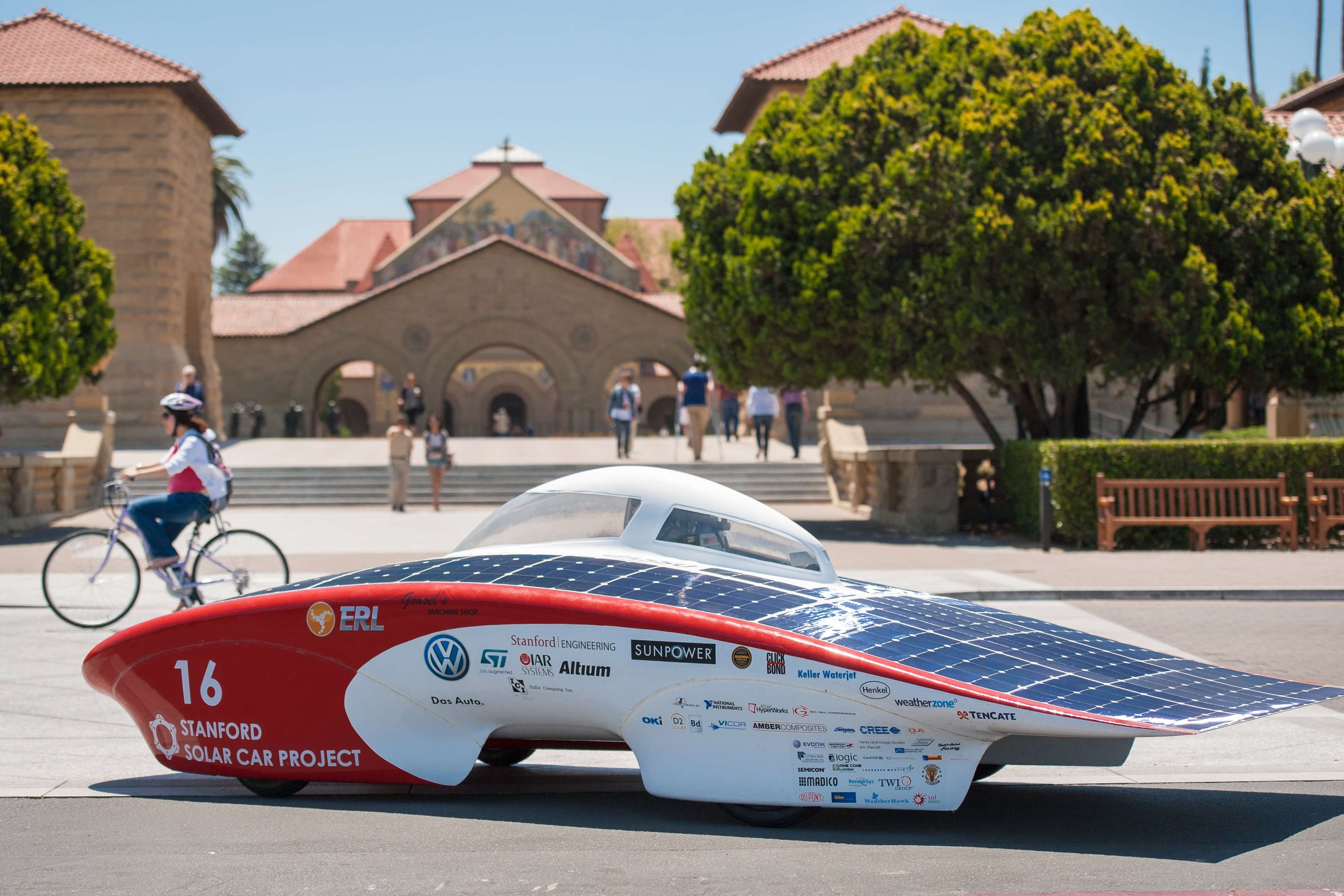 A solar-powered vehicle stands parked outside a building with a person riding a bike in the background.