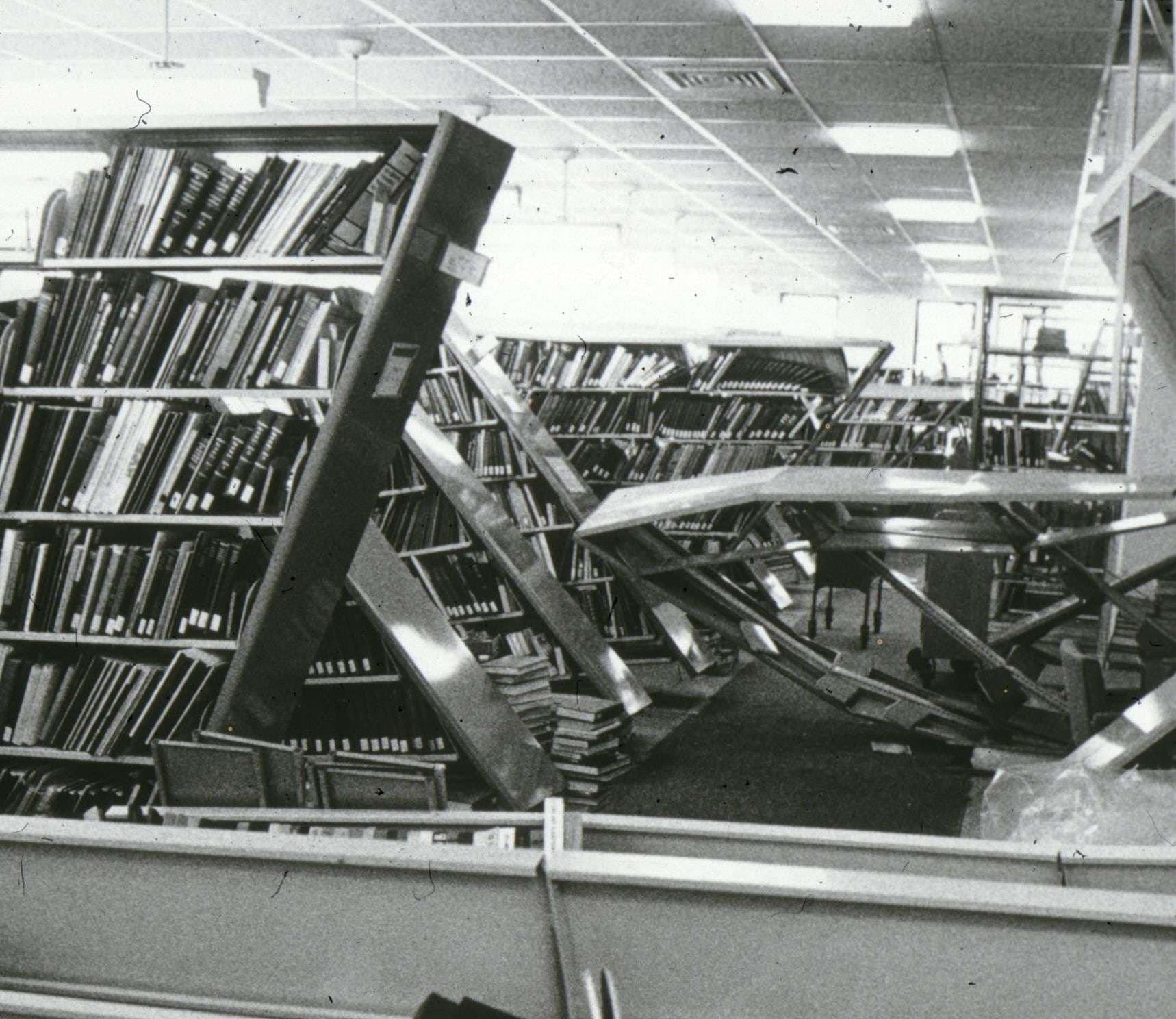 A black and white photo of a library featuring fallen bookshelves and books scattered on the floor.