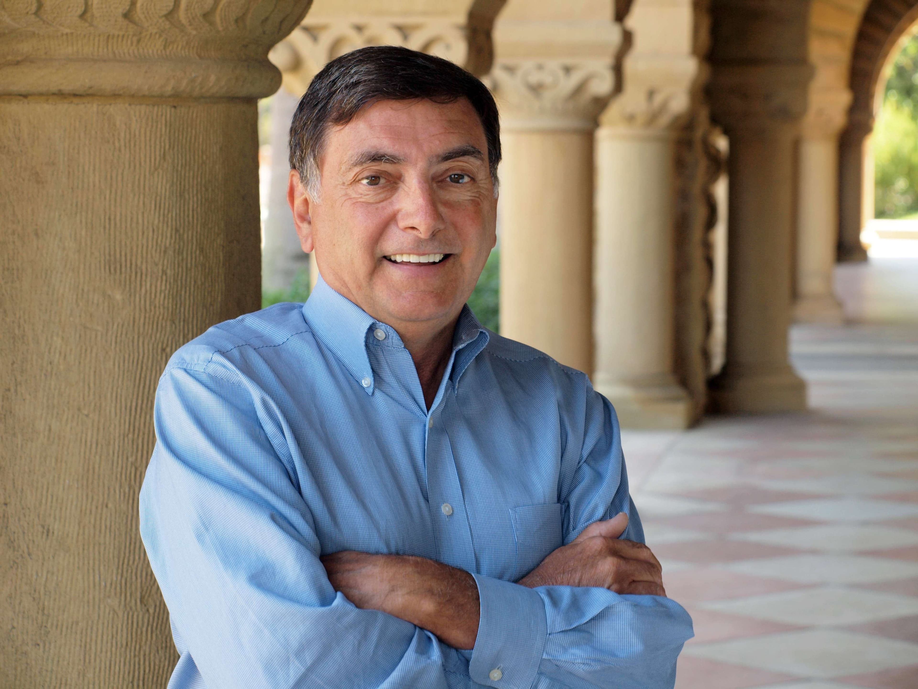 A man in a blue shirt stands confidently in front of tall and elegant pillars.