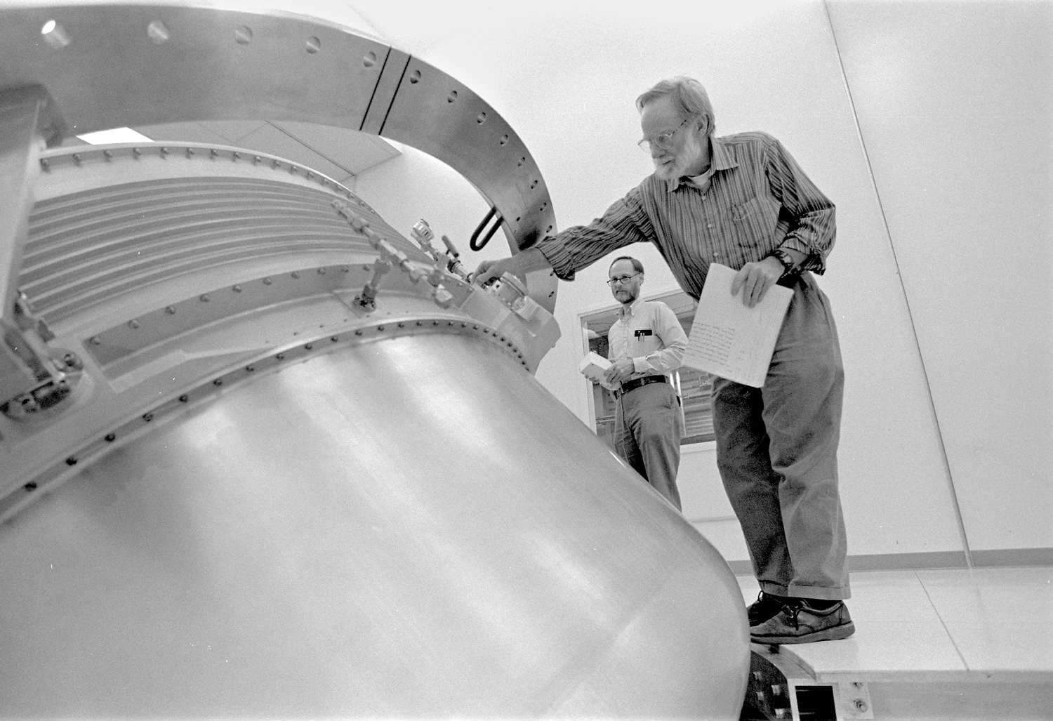 A man inspects a large metal object, accompanied by another man who watches nearby.