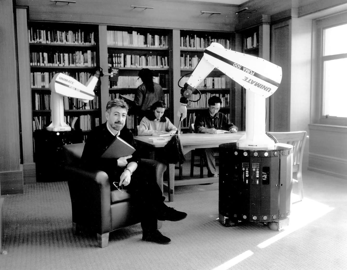 A man poses in a chair sitting next to a robot dusting the table, while three individuals work in the background.