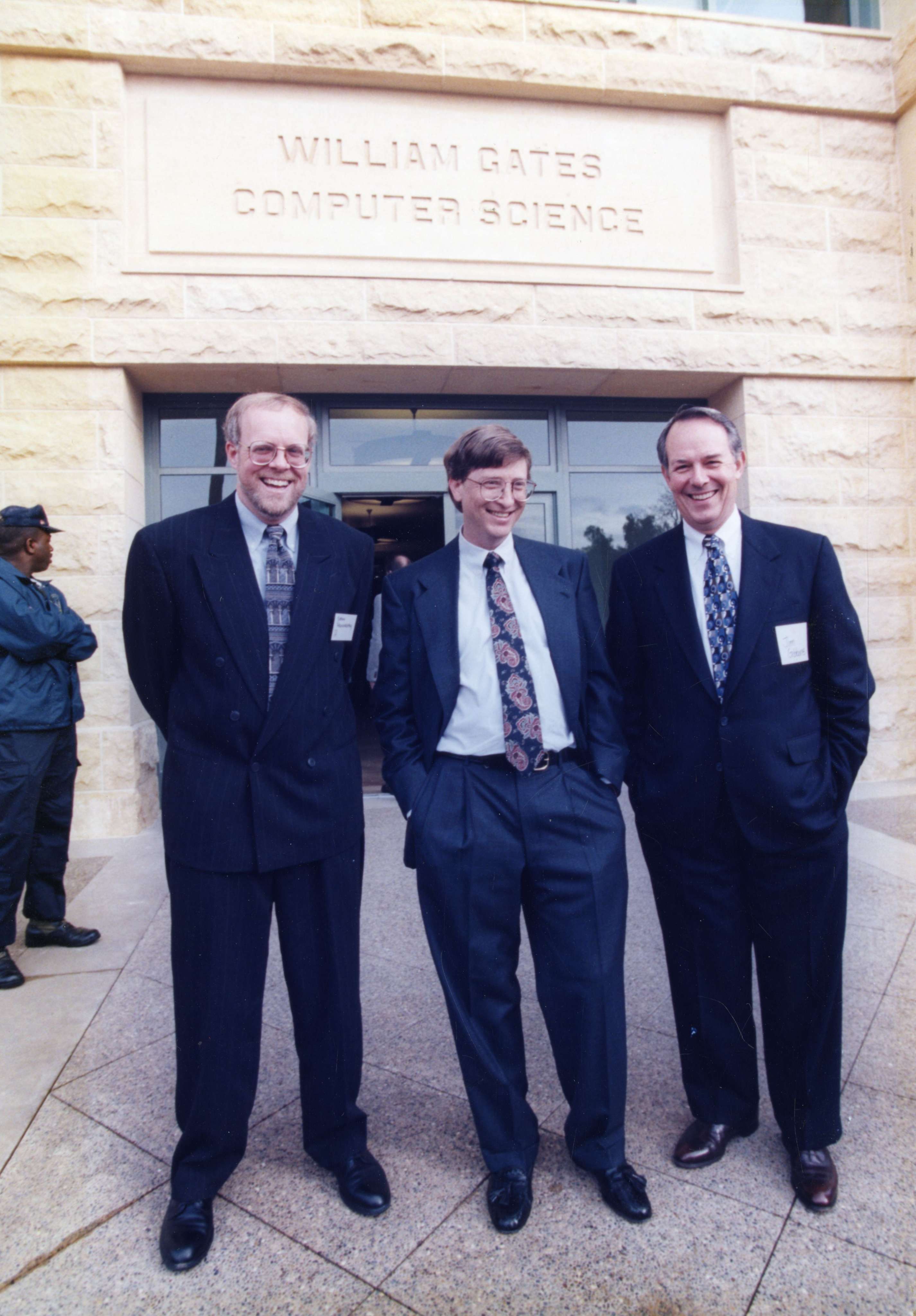 Three men in formal suits standing together outside the William Gates Computer Science building.