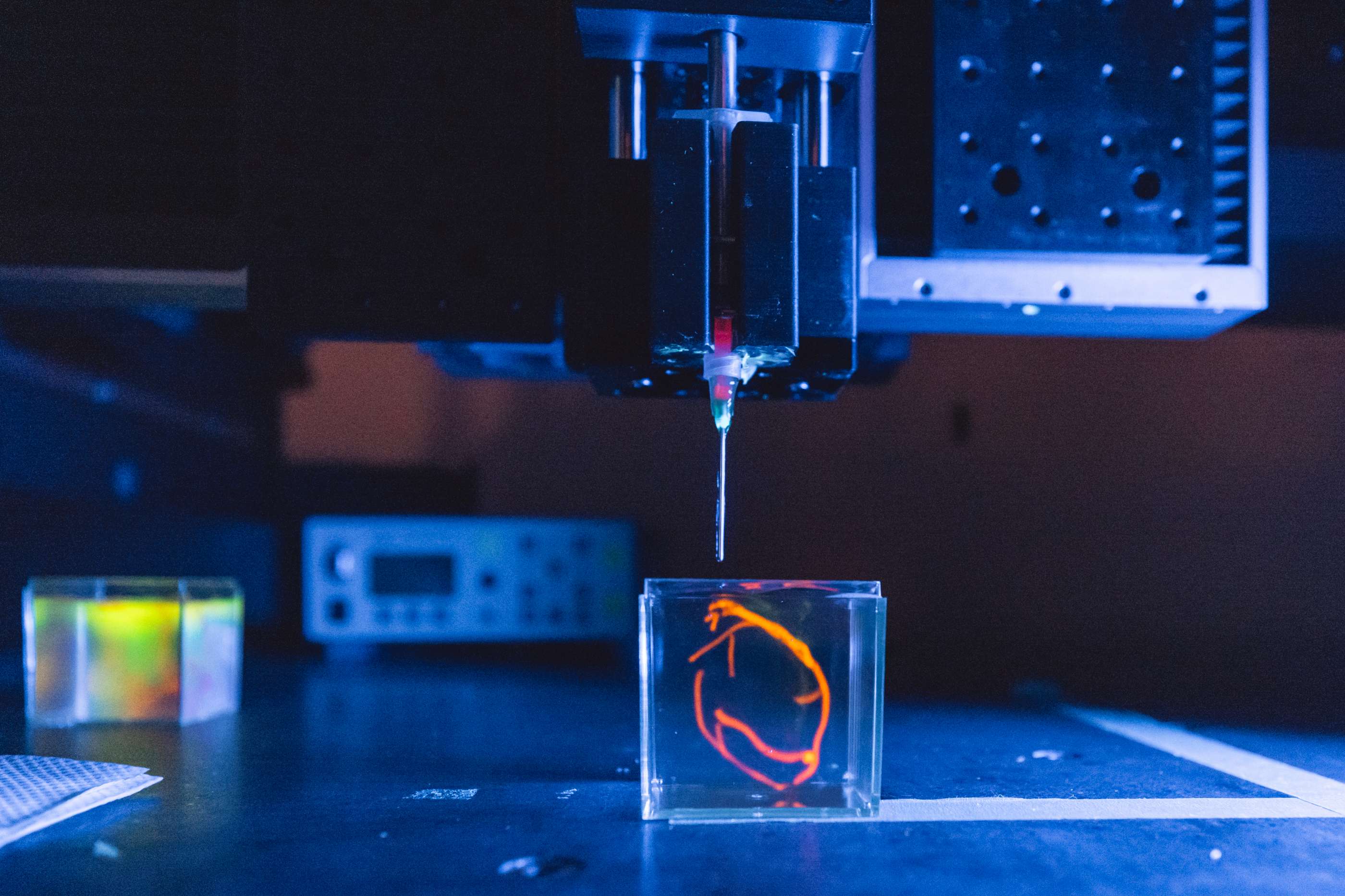 An injection needle extends below the arm of a 3D printer and hovers over a clear block with orange vein-like contents.