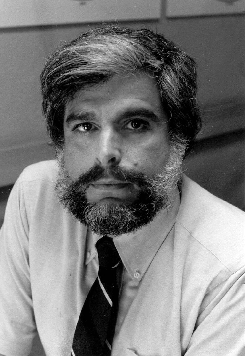 A black and white image of a man with a full beard, wearing a tie, sitting at a desk.