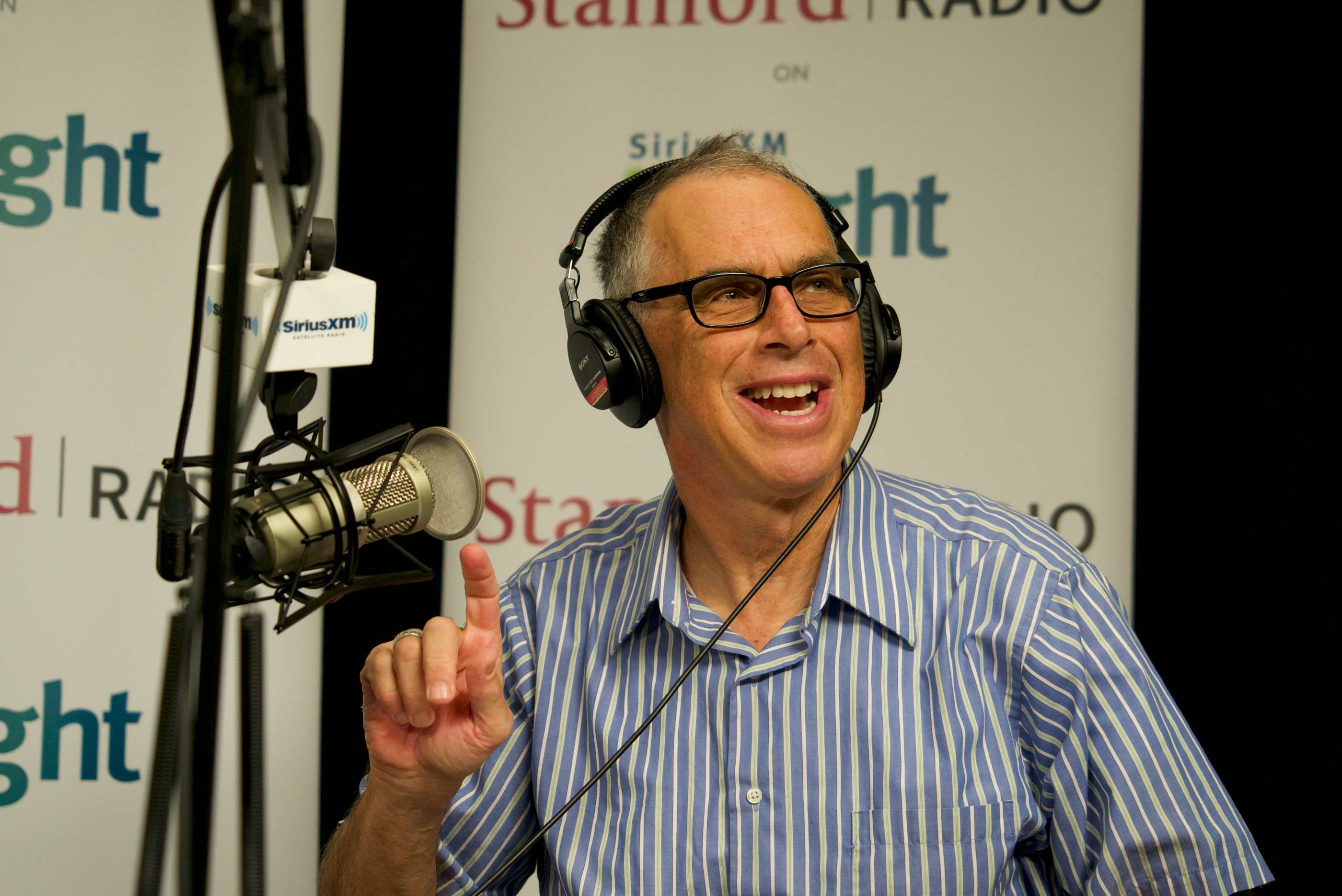 A man with glasses and headphones speaks enthusiastically into a microphone.