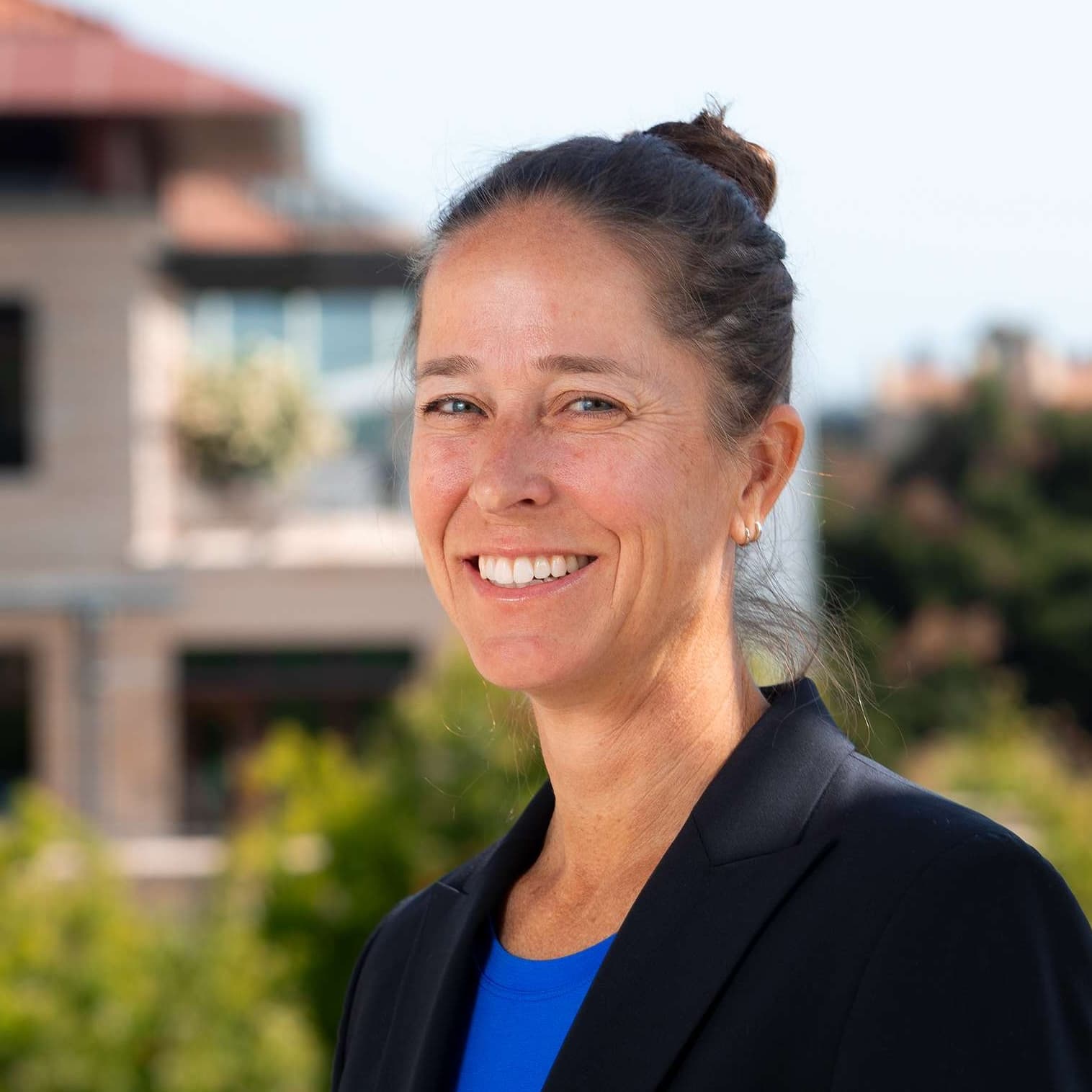 A woman with dark hair in a bun smiles warmly, wearing a blue shirt and black blazer.