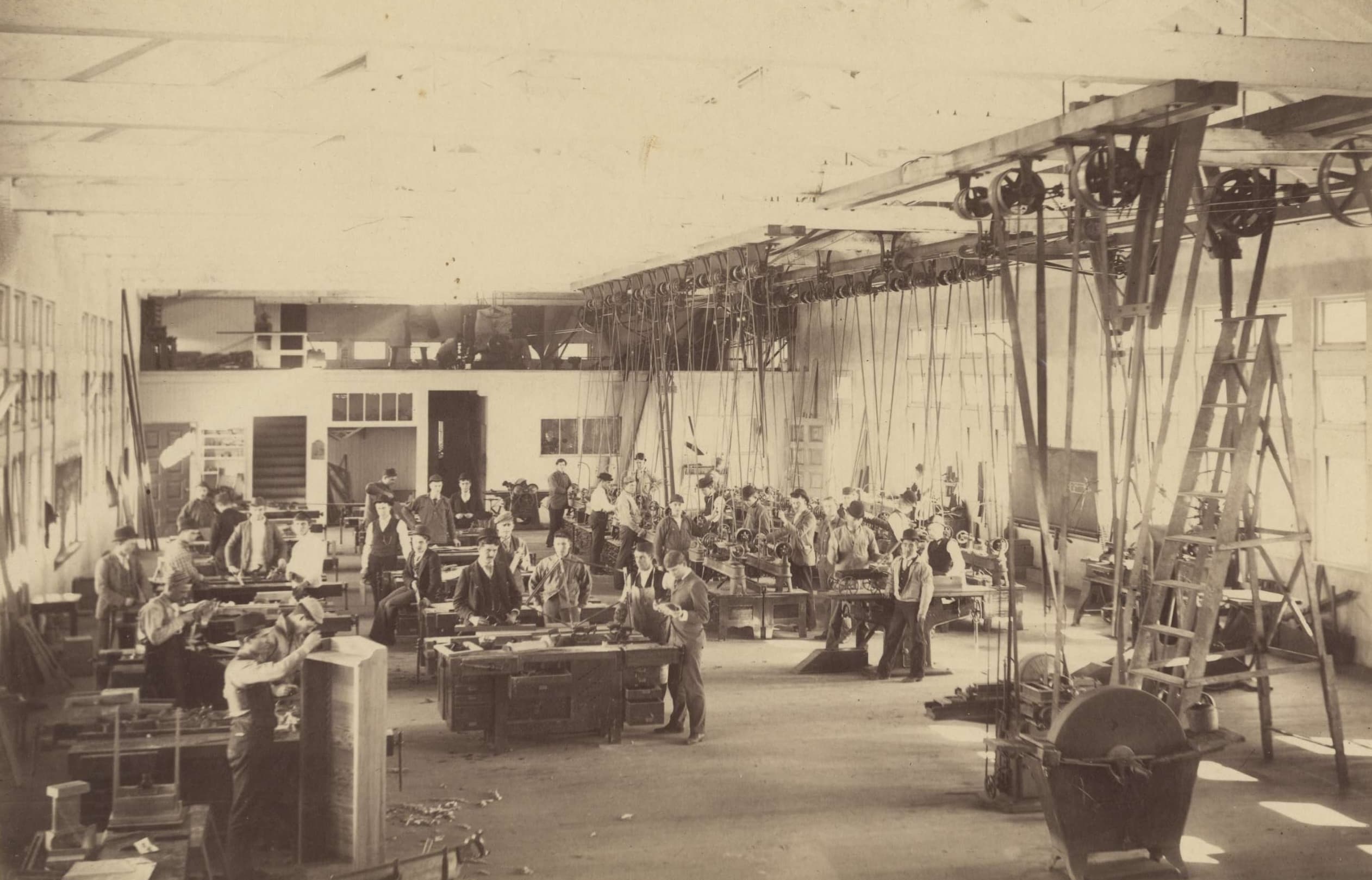 Old photograph depicting a busy carpentry workshop, featuring men in aprons engaged in work under bright windows.