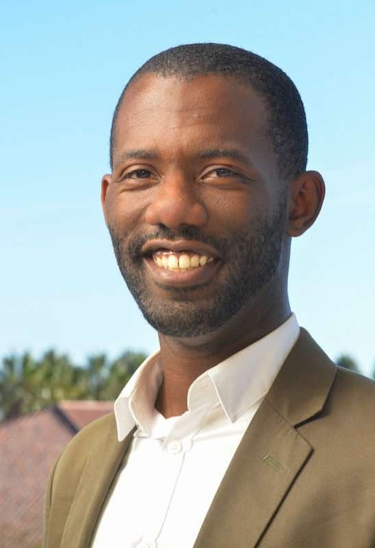 Smiling man in a brown blazer stands outdoors.