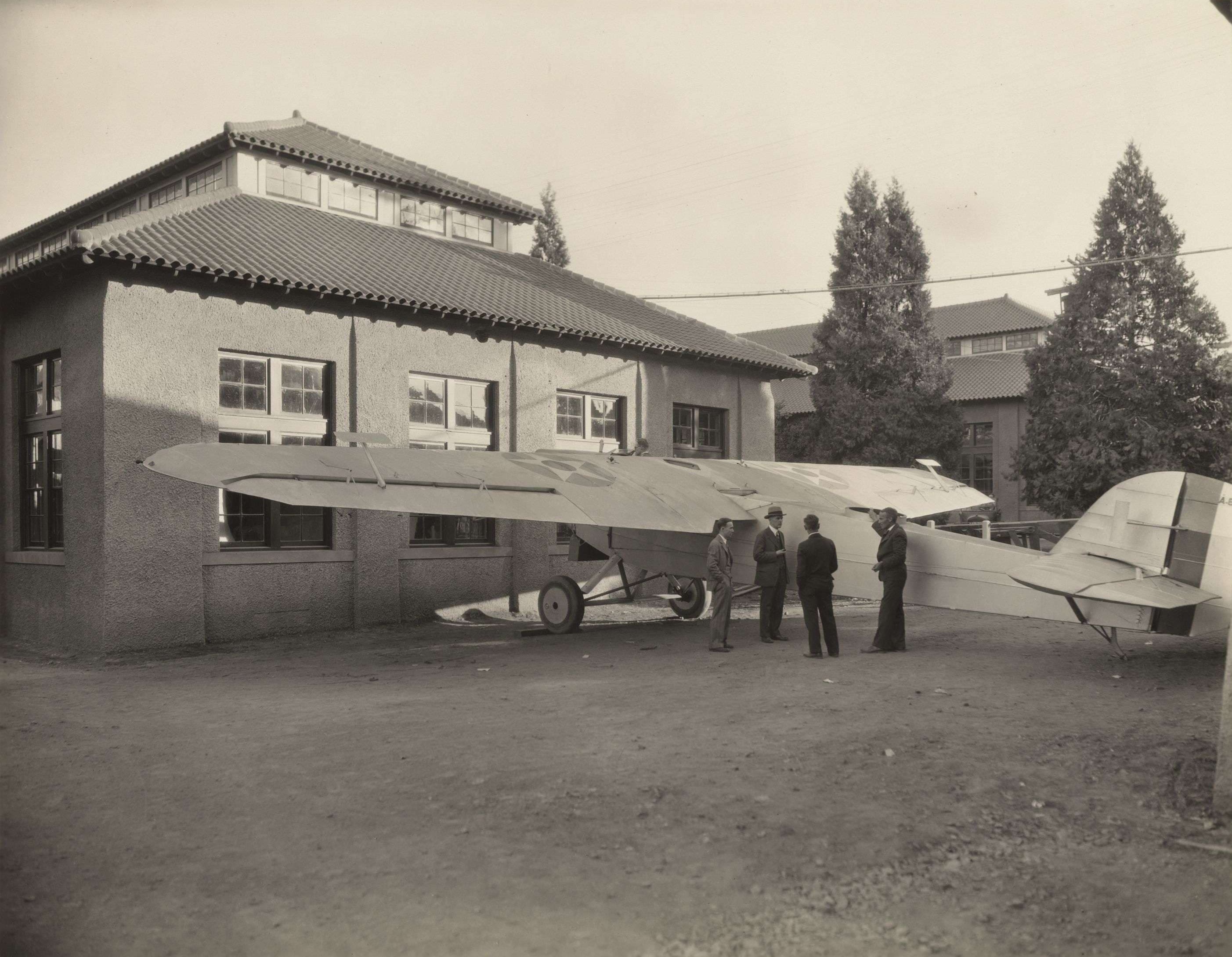 Showcasing a plane parked in front of a historic building, with a group of men standing by, capturing a moment from aviation history.