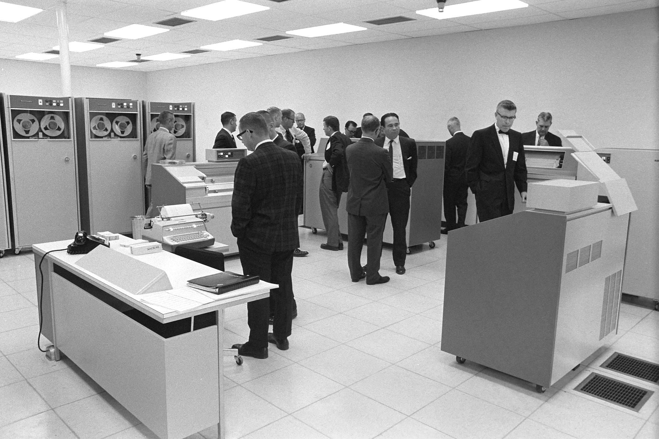 A room filled with several men gathered around a couple of computers, engaged in discussion and collaboration.
