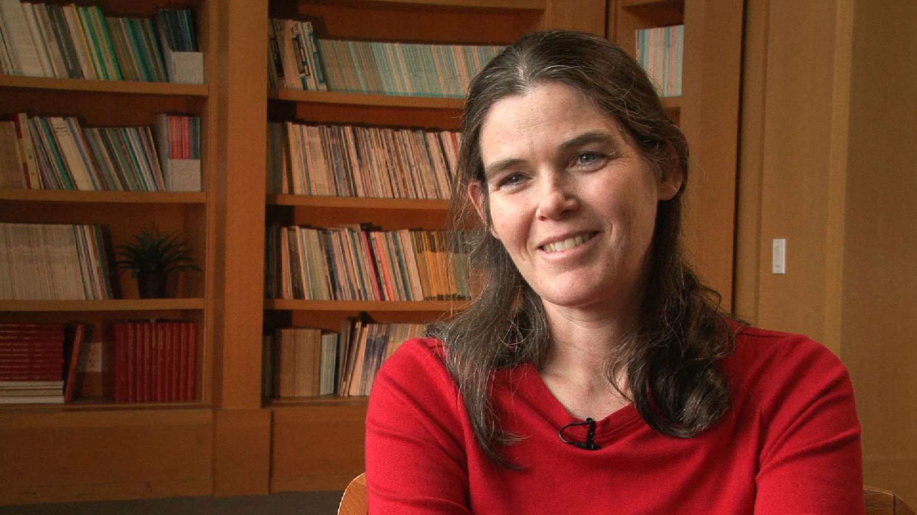 A woman with long brown hair wearing a red sweater sits and smiles in front of bookshelves filled with books.