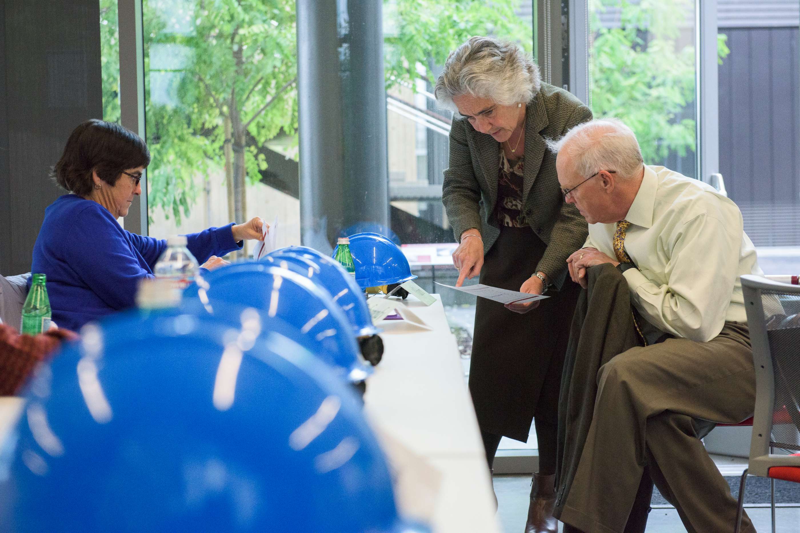 A woman shows a document to a man while another woman reads paperwork, surrounded by blue safety helmets on a table.